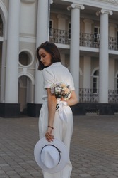 Back view of young elegant bride in white blouse and pants stranding with flower bouquet and hat in hands behind back lit by sunlight against historic building  