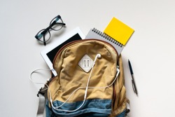 Hipster brown-blue textile backpack, full of school supplies, blank screen tablet, earphones, glasses, notebook, yellow stickers on white background. Back to school concept. Close up, copy space, flat
