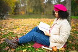 Serious dark-skinned intelligent african american woman, beautiful stylish ethnic girl reader reading a book in a golden autumn park in autumn, sitting on the grass