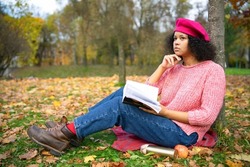 Serious dark-skinned intelligent african american woman, beautiful, pensive ethnic reader, daydreaming near a book in a golden autumn park in autumn, sitting on the grass