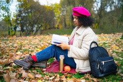 Serious dark-skinned intelligent African american woman, beautiful stylish ethnic girl reader reading a book in a golden autumn park in autumn, sitting on the grass