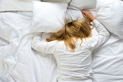 A woman covers her face with a pillow. The woman lies tired on a white bed. Oversleep, not getting enough sleep concept. Young beautiful blonde woman lying in bed. Early wake up concept