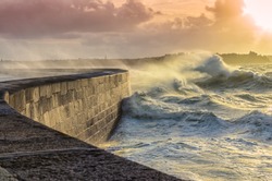Big waves crushing on curved stone pier, on stormy weather with vivid sunset, big tide, Saint Malo, France.