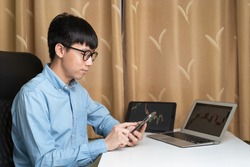 Happy young Asian businessman or investor using smartphone for trading, investing on new cryptocurrency or stock trading platform at laptop, tablet with stock tickers and graphs. Young money concept.