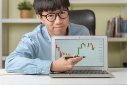 Happy young Asian businessman or investor pointing on trading tickers or graphs on laptop to teach online investing courses on stock market or Cryptocurrency as Bitcoin at home. Young money concept.