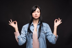Attractive caucasian brunette girl in shirt in lotus position showing zen and mudra gesture isolated on black studio background.