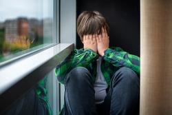A frustrated, sad and lonely teenager of 12 years old is sitting on the windowsill of the house, covering his face with his hands. Children's social psychology and emotions.