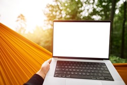 Design mock-up of a modern laptop computer with a blank white screen. Hiker is resting in a hammock in the woods. Using computer outdoors concept.