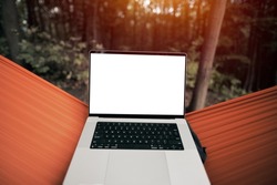 Design mock-up of a modern laptop computer with a blank white screen. Hiker is resting in a hammock in the woods. Using computer outdoors concept.