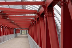 Pedestrian crossing, construction of red metal structures. The roof is made of steel channels connected to each other. Red iron beams on bolts and rivets.