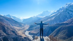 Man wearing a beanie and blue jumper, spreads his arms wide, breathing deeply the fresh mountain air. His gesture represents freedom and happiness. Below a long valley stretches in Himalayas.