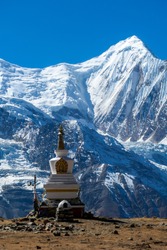 A stupa with Annapurna Chain as a backdrop, Annapurna Circuit Trek, Himalayas, Nepal. High mountains covered with snow. Land in front of the stupa is barren and dry. Some prayer's flag next to it.