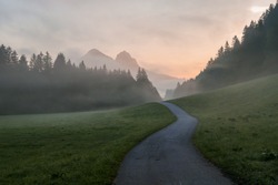 Early morning haze in the Alps. There is a narrow road leading to high mountains through a meadow. The high Alpine peaks are shrouded with light fog. The sky is turning pink. Daybreak. Calmness