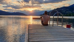 A man in full cup sitting at the end of a wooden pier of Millstaetter lake and enjoys the sunset. The sun sets behind high Alps. Calm surface of the lake reflects the orange sky and the mountains