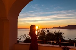 Woman with champagne glass in dress with scenic sea view at sunset from luxury apartment on Mediterranean Sea. Terrace balcony with arch at coastline in Praiano, Amalfi Coast, Campania, Italy, Europe