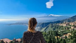 Tourist woman with panoramic view on snow capped Mount Etna volcano and the Mediterranean coastline on a sunny day seen from ancient Greek theater of Taormina, island Sicily, Italy, Europe, EU. Awe