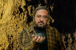 A geologist man in clean clothes with glasses holds in his hand a piece of coal on a blurred cave background. Illustration of research on fossil fuels with a non-renewable resource.