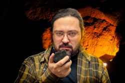 A geologist man in clean clothes with glasses holds in his hand a piece of coal on a blurred cave background. Illustration of research on fossil fuels with a non-renewable resource.