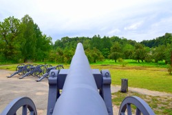 A massive cannon seen from the loading side with two wheels on the sides and three more cannons visible in the distance located on a path near a lawn and a set of trees seen during autumn day