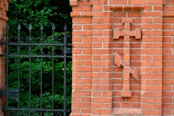 A close up on the symbol of an orthodox church inside a frame made out of red bricks and signifying an old cemetery located next to a metal gate and a small garden seen in Poland 
