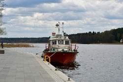 A view of a luxirious boat parked next to a small wooden marina, pier or platform with a metal ladder for entering and exiting the vessel seen next to a lush forest or moor on a cloudy spring day
