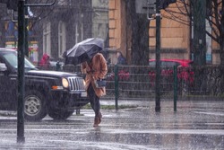 A woman in a brown coat under an umbrella crosses a busy street in heavy rain. Heavy precipitation in the city. Flooding of city streets.