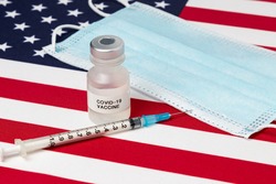 Closeup of vial with Covid-19 vaccine, syringe and needle, and face mask on United States of America flag. Concept of medical research and treatment, coronavirus pandemic and healthcare.