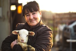 Farmer woman carrying lamb in arms smiling sunset day