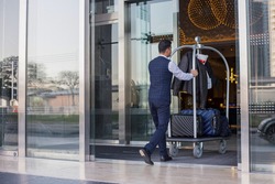 Young man pushes luggage cart with suitcases, bags and backpacks to the entrance. Metall baggage trailer with luggage