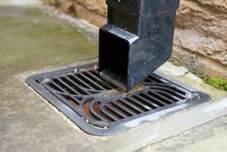 Square black plastic drainage downpipe with a down spout shoe at the end of it. Fixed to a brick wall. Leading to a black plastic square gulley grid type drainage cover. Day outdoors. Close up.