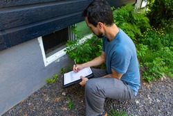 Indoor damp & air quality (IAQ) testing. A close up view on environmental home quality inspector at work, filling in a form as he inspects the exterior of a cellar window, with room for copy.