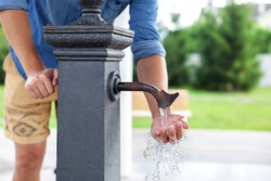 Man washing his hand in faucet water. Сity water tap with drinkable water in park. Drinking column. Ancient black column for distribution of drinking water installed on street. Watertap on hydrant 
