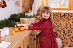 Smiling little girl in dress cooking Christmas biscuits at home. Happy child sits on chair next to Xmas cake in kitchen in New Year decorations with Christmas tree, gift. Kid in kitchen on Xmas eve