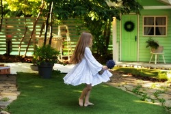 Little girl is spinning barefoot on ground in white dress in summer garden. Beautiful young girl spinning in dance and enjoying nature. Kid walking barefoot on green grass and spin in backyard at home