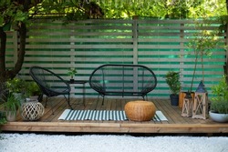 Modern lounge outdoors in backyard. Terrace house with plants, wooden wall and table, comfortable sofa, armchair and lanterns. Wooden verande with garden furniture. Cozy space in patio or balcony. 
