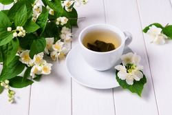 Green tea with jasmine in a white cup on a white wooden background and a branch of blooming jasmine