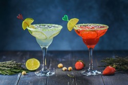 Two margarita cocktails with decorations on craft blue background with berries, nuts and greens