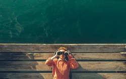 Young Beautiful Girl Lies On A Pier Near The Sea And Looks Through Binoculars On Tje Sky. Travel Search Journey Concept