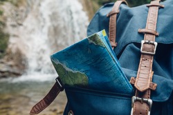 Hipster Blue Backpack, And Map Closeup. View From Front Tourist Traveler Bag On Waterfall Background. Exploring Adventure Hiking Concept