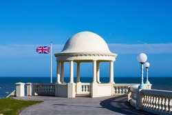 Rotunda on Bexhill seafront overlooking the ocean with the British flag flying in the background and balustrade and lamp posts in the foreground on a summer day with clear blue sky