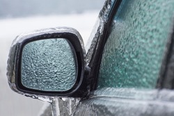 Ice-covered car mirror with icicles, glass and side doors. Icing vehicle on a winter day after freezing rain in Ukraine. Blurred background.