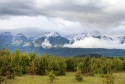 Beautiful mountain landscape with snowy peaks of the Eastern Sayan Mountains and green meadows of the foothill Tunka valley with low clouds on a rainy day. Travel to the highlands. Natural background