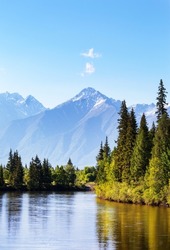 Beautiful summer landscape with green forest at the riverside and mountain snowy peaks on sunny June day. Siberia, Baikal region, Eastern Sayan Mountains,  Buryatia, Tunka foothill valley, Irkut River