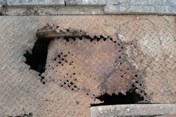 The manhole cover is made of brown rusted sheet steel. Corrosion from sunlight or water and chemical oxidation (combination of steel surfaces, moisture and oxidation) can be detrimental to passers-by.