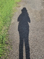 Looking down at the shadow of a woman on a concrete road, to the left is a green lawn. In shadow she was wearing a hat and can see long legs. Sunlight shines through the body, creating funny shadows.