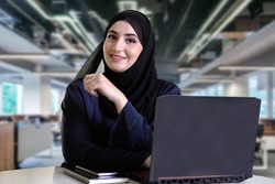 Happy Emirati woman on Abaya lookikng at the front camera with computer laptop, notebook and blurred office