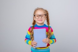 cute little girl in a multicolored sweater and glasses holds a book on a white background , smiling

