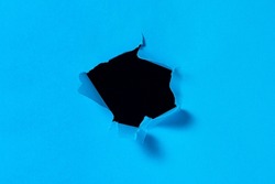 A hole in blue paper with torn edges and a black isolated background inside. Ripped blue paper, copy space