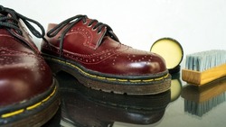 Maroon wingtip brogue made of genuine leather side by side with brush and shoe polish on white background