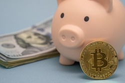 Pink piggy bank, stuck of dollars and bitcoin coin on a blue background. The future of virtual money currency, account security, investment in cryptocurrency.